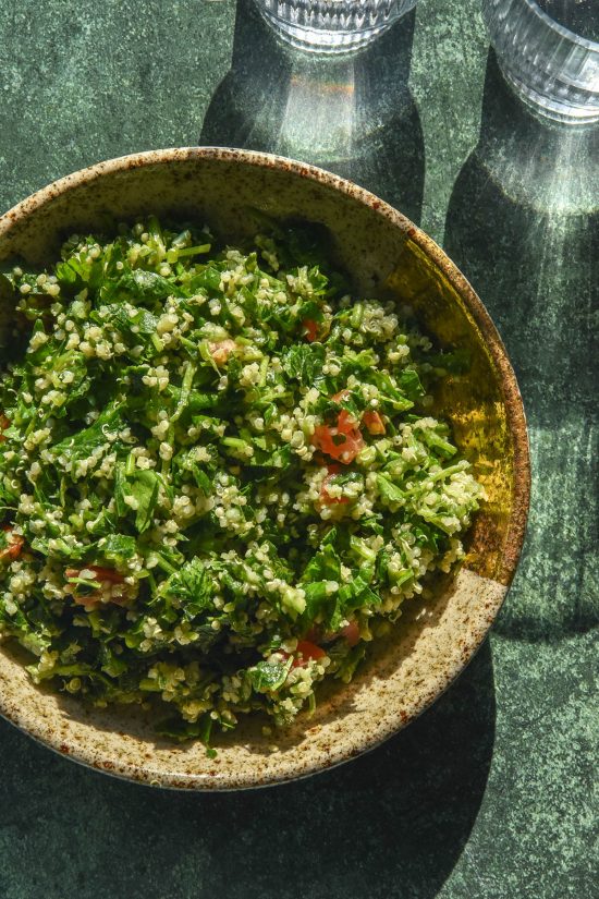 An aerial image of a grey speckled ceramic bowl filled with low FODMAP tabbouleh on an olive green backdrop. Two sunlit glasses of water sit to the top right of the image.