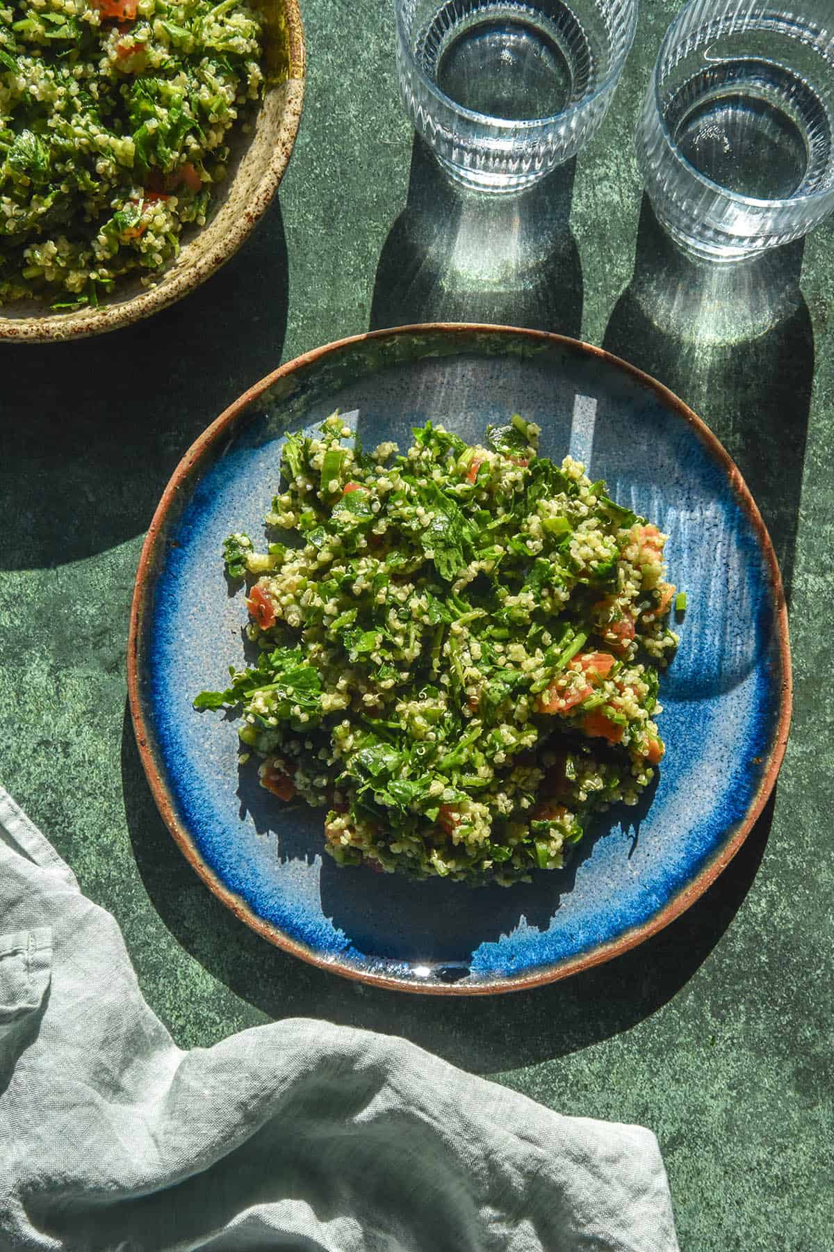 An aerial image of a bright blue ceramic plate topped with low FODMAP tabbouleh. The plate sits atop an olive green backdrop in bright sunlight. Two glasses of water sit in the top right of the image, while another bowl of tabbouleh sits in the top left.