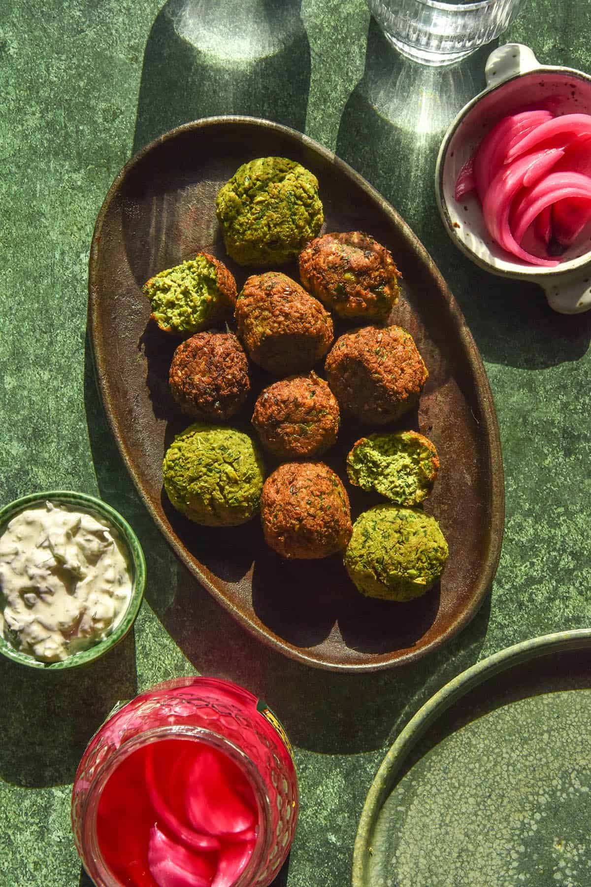 An aerial image of low FODMAP falafel on a dark grey platter atop an olive green backdrop. The falafel are surrounded by a small plate of low FODMAP tzatziki, two bowls of pickled red onions and a two sunlit glasses of water.