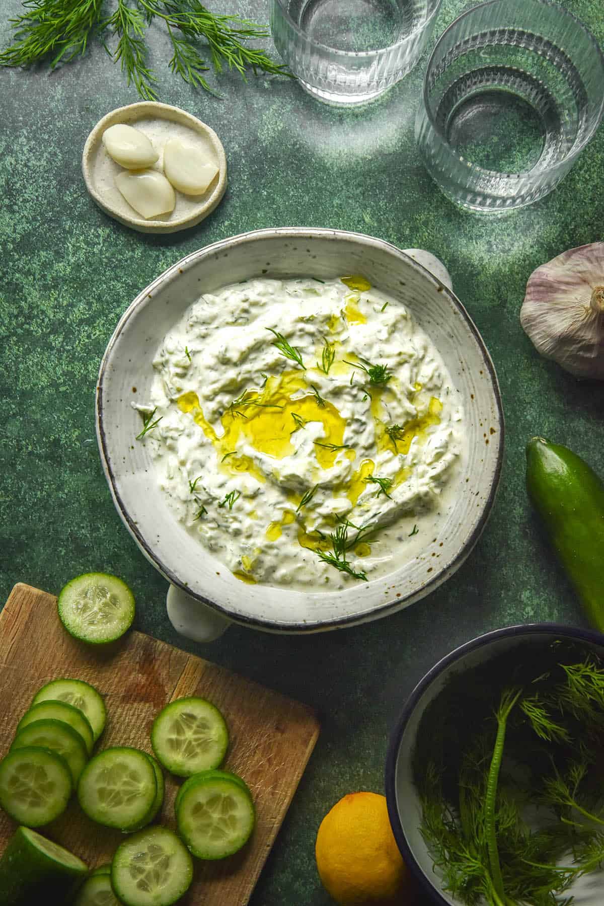 An aerial image of a bowl of low FODMAP tzatziki on an olive green backdrop. The bowl is surrounded by ingredients used to make the tzatziki and two sunlit water glasses. 