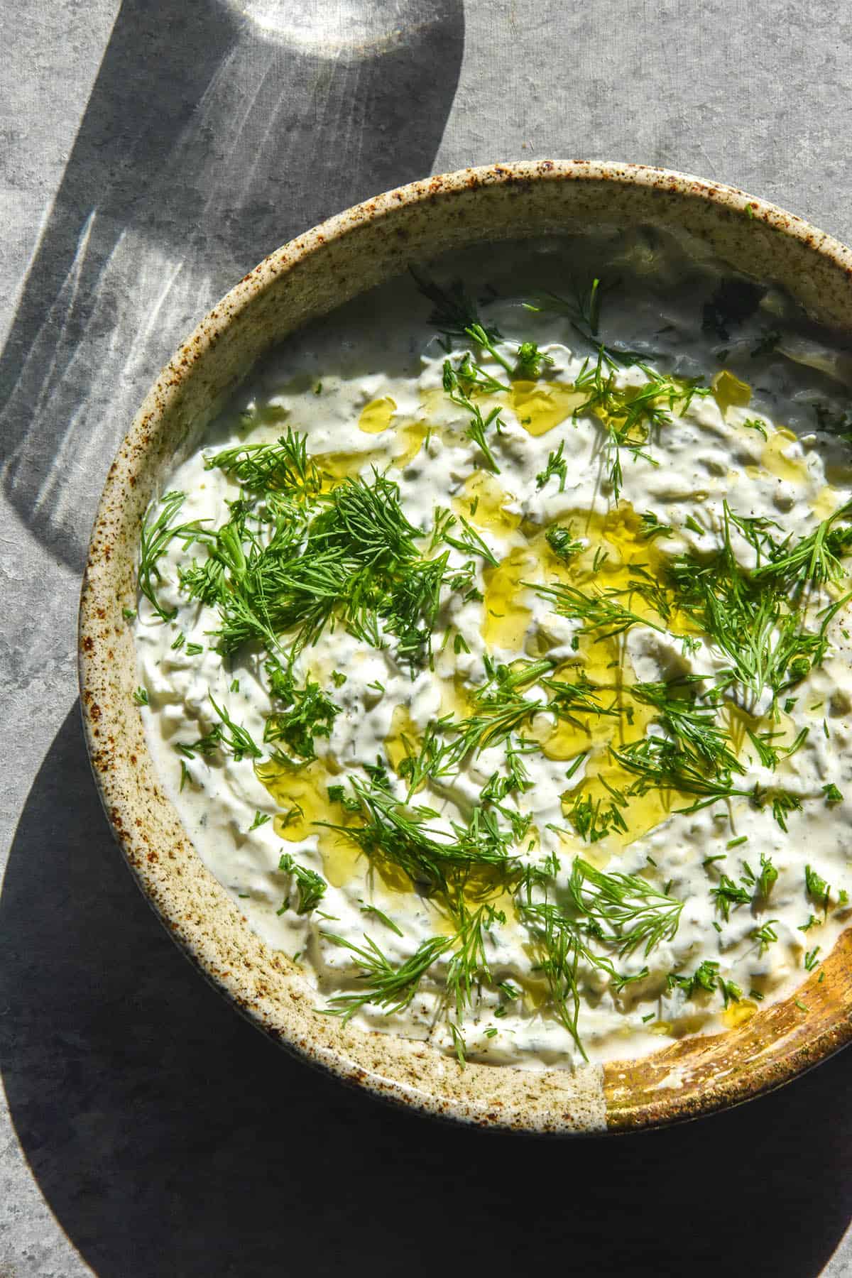 An aerial image of a beige ceramic bowl filled with low FODMAP tzatziki. The tzatziki is topped with plenty of dill and olive oil. It sits atop a light grey concrete background and a sunlit glass of water sits to the top left of the image.