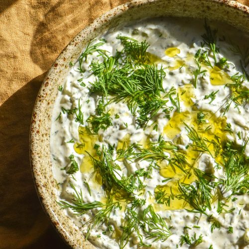 An aerial image of a beige ceramic bowl filled with low FODMAP tzatziki. The tzatziki is topped with plenty of dill and olive oil. It sits atop a mustard coloured linen tablecloth and a sunlit glass of water sits to the top right of the image.