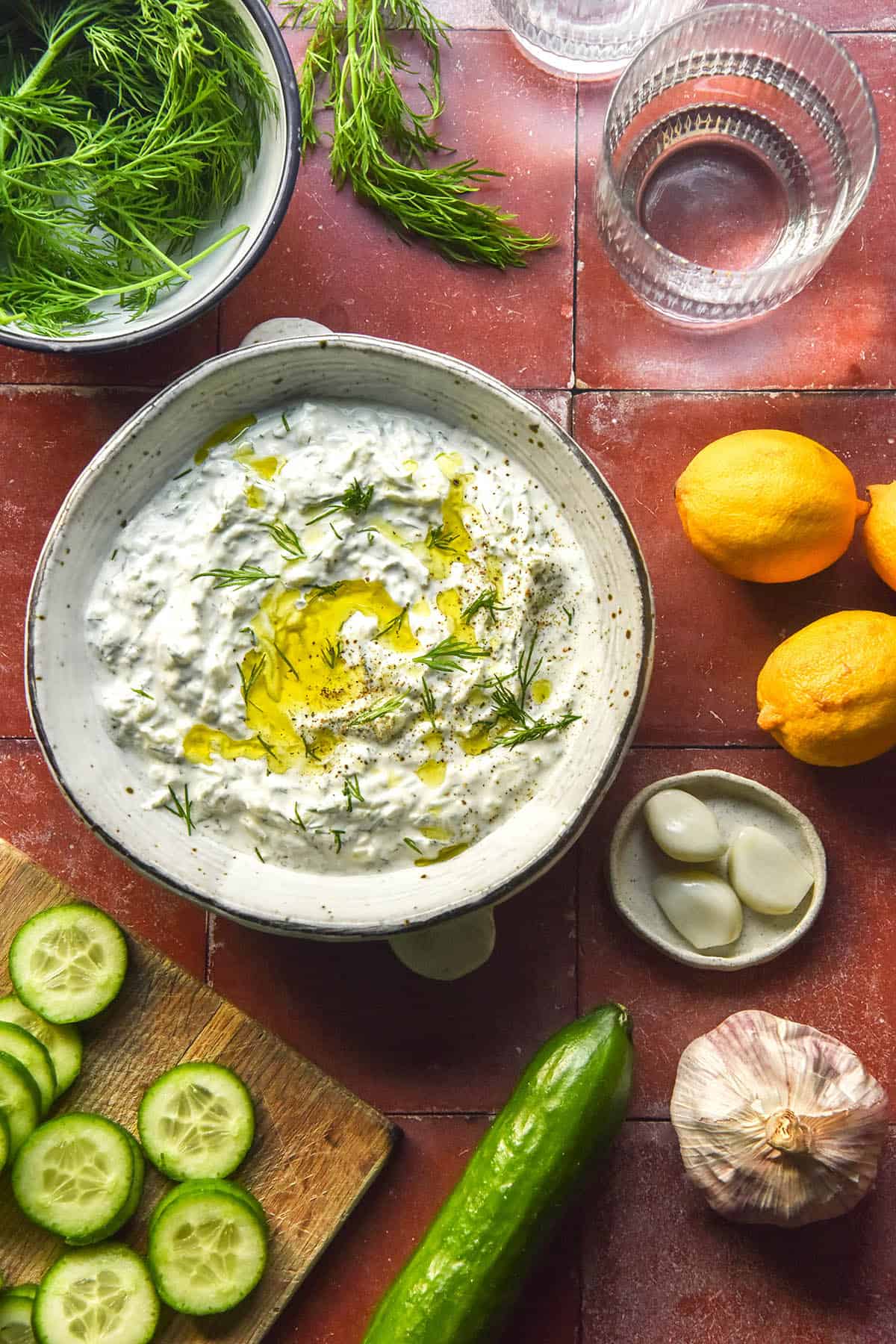 An aerial image of a white ceramic bowl filled with low FODMAP tzatziki on a terracotta tile backdrop. The tzatziki is surrounded by the ingredients used to make the dip as well as two sunlit glasses of water in the top right corner.
