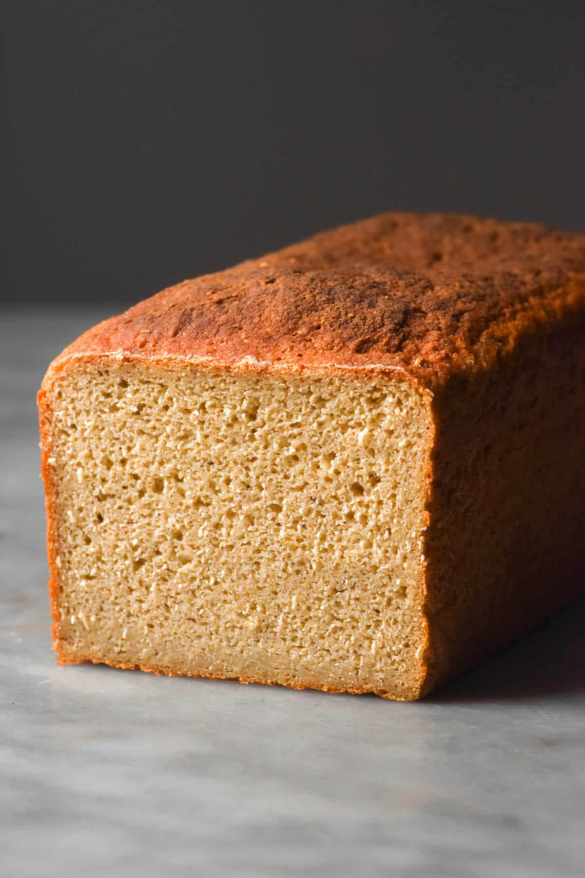 A side on image of a loaf go gluten free quinoa bread that has been sliced to reveal the inner crumb. The loaf sits atop a white marble table against a dark backdrop