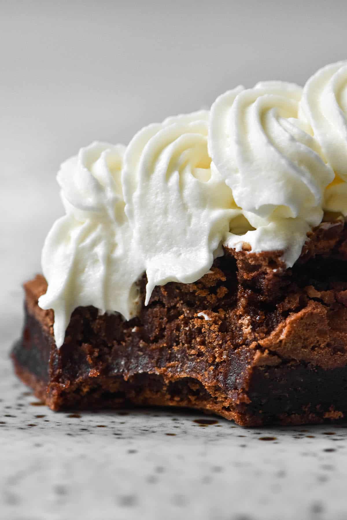 A macro side image of a brownie topped with piped lactose free whipping cream. The brownie has a bite taken out of the from left corner and sits on a white speckled ceramic plate against a white background