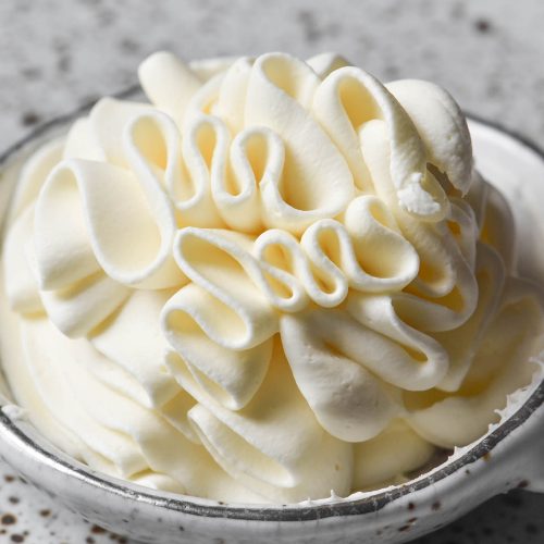 A side on image of a small white ceramic bowl filled with piped lactose free whipping cream. The bowl sits atop a white speckled ceramic plate