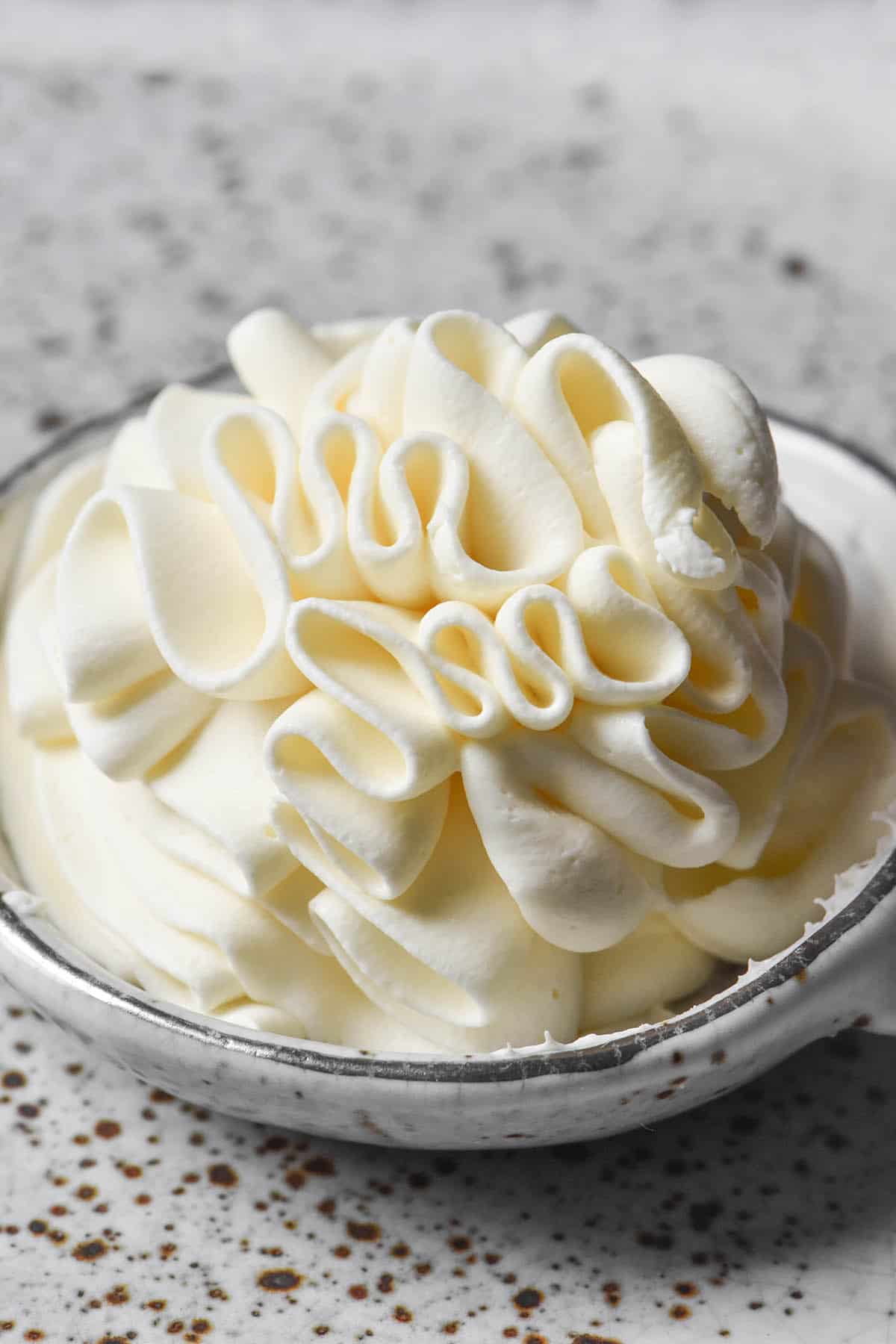 A side on image of a small white ceramic bowl filled with piped lactose free whipping cream. The bowl sits atop a white speckled ceramic plate