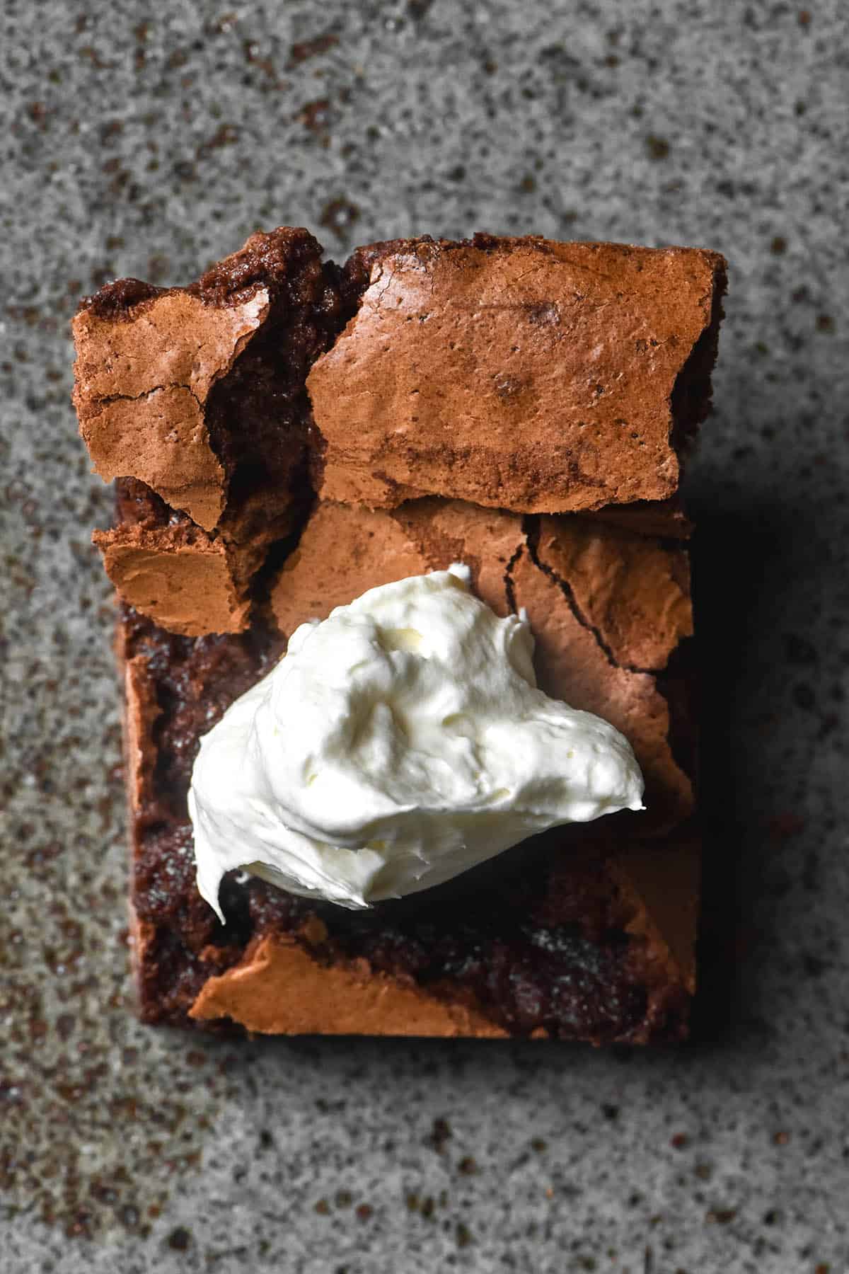 An aerial image of a brownie topped with lactose free whipped cream on a dark grey speckled ceramic plate