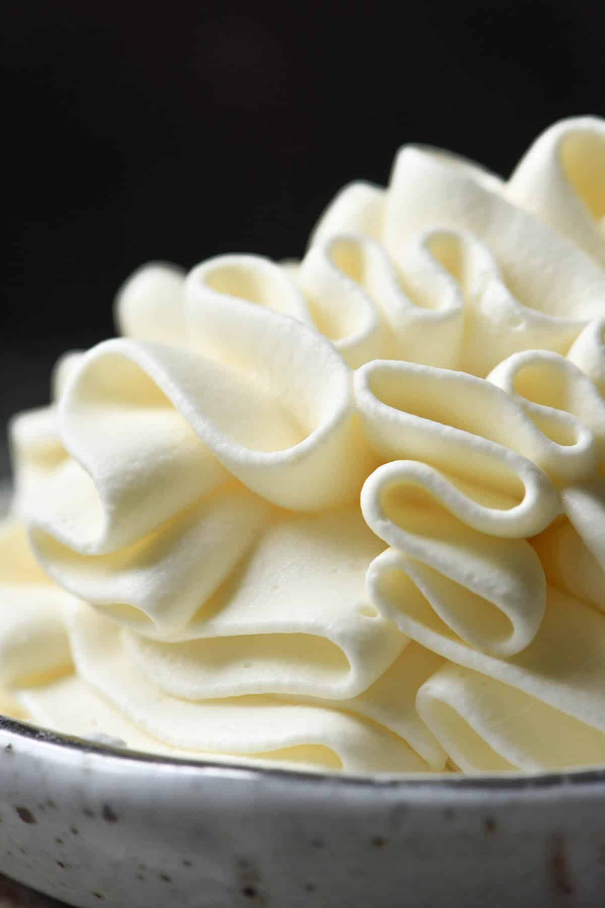 A side on macro image of lactose free whipping cream piped into a small white ceramic bowl against a dark backdrop