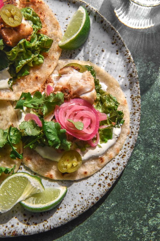 An aerial image of a low FODMAP fish taco topped with crema, pickled jalapeños, coriander and pickled red onion. The tacos sit on a white speckled ceramic plate atop a medium green backdrop. A sunlit glass of water sits in the top right corner