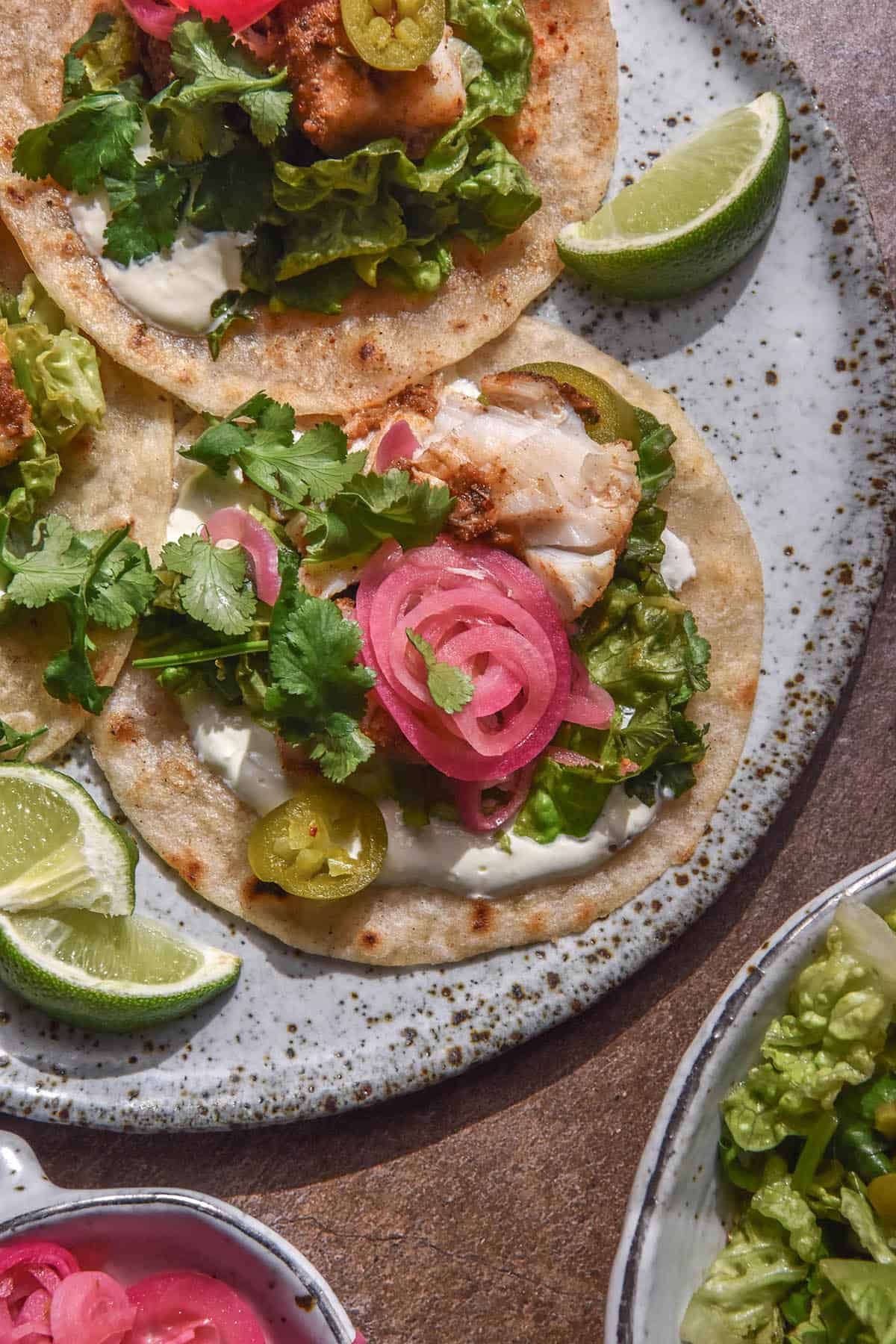 An aerial image of a low FODMAP fish taco topped with crema, pickled jalapeños, coriander and pickled red onion. The tacos sit on a speckled white ceramic plate atop a medium brown backdrop. The plate of tacos is surrounded by smaller bowls with topping ingredients.