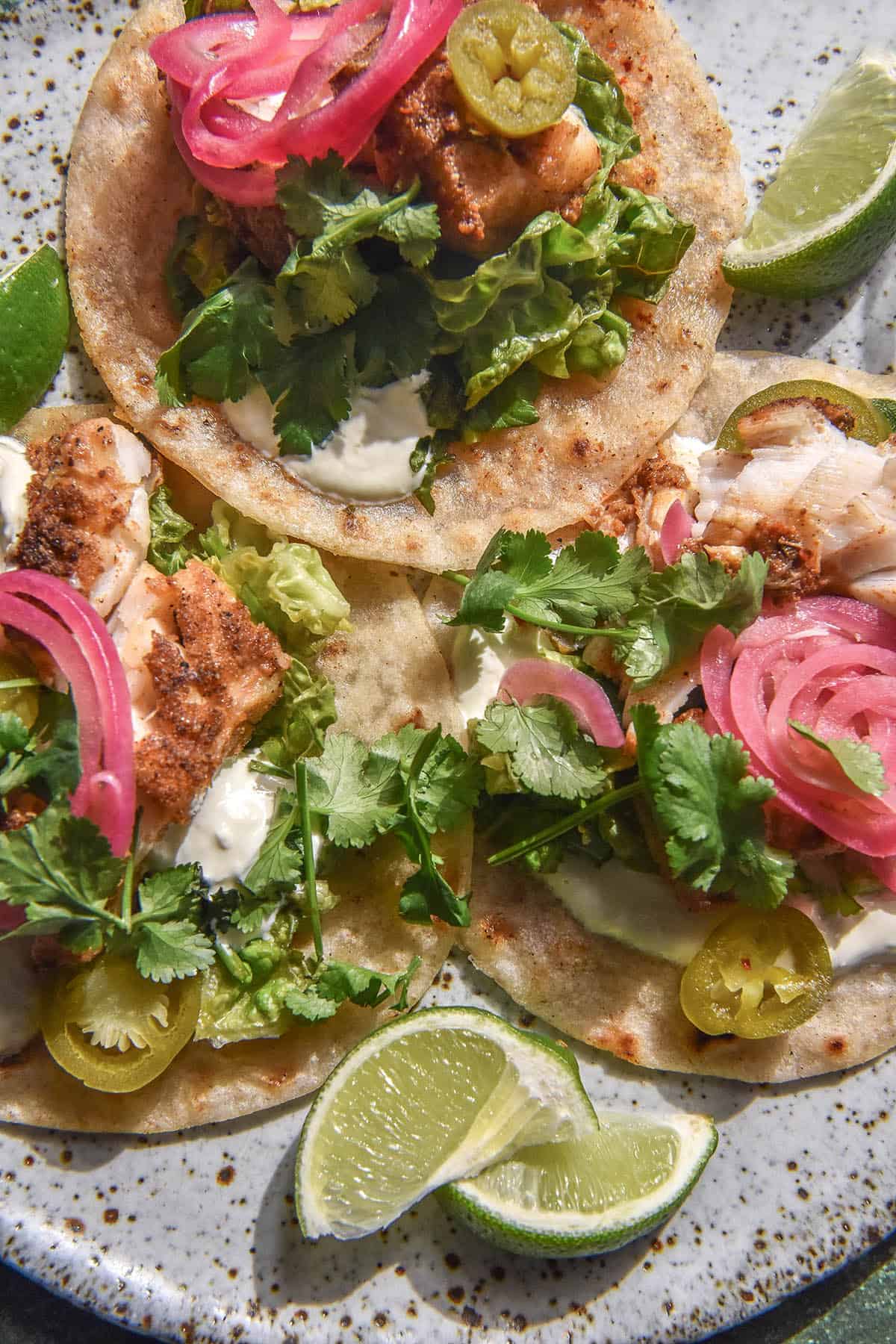 An aerial image of three low FODMAP fish tacos on a white speckled ceramic plate. The tacos are topped with grilled spiced fish, crema, lettuce coriander mix, pickled jalapeños, pickled red onion and coriander. They are surrounded by wedges of lime.