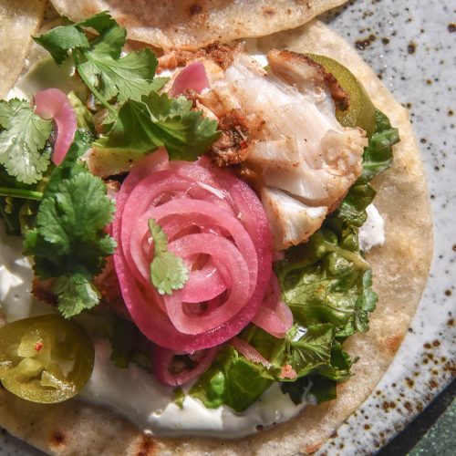 An aerial close up image of a low FODMAP fish taco topped with low FODMAP pickled onions, coriander and pickled jalapeño. The tacos sit atop a white speckled ceramic plate against a medium grey backdrop