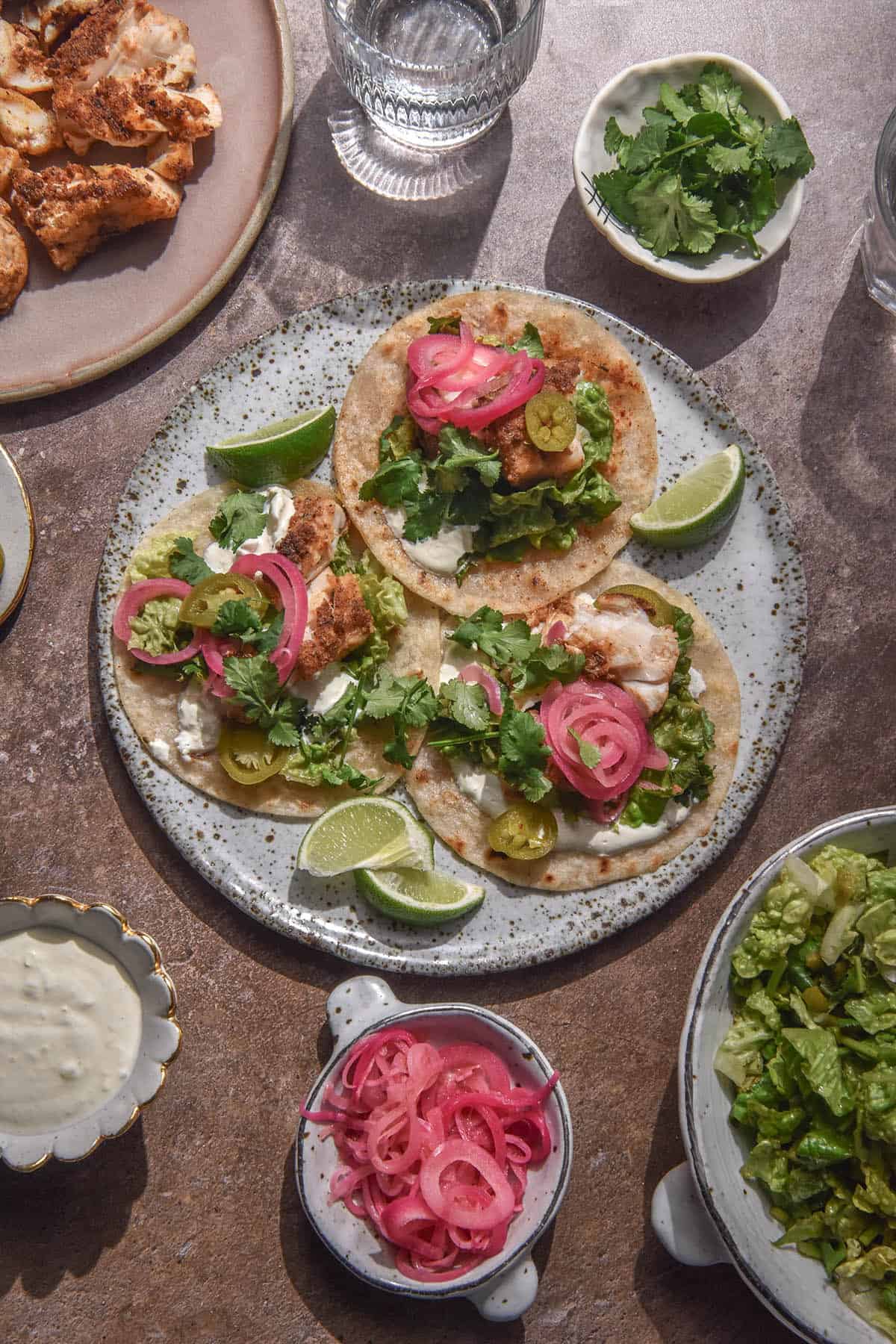 An aerial image of three low FODMAP fish tacos on a white speckled ceramic plate. The tacos are topped with grilled spiced fish, crema, lettuce coriander mix, pickled jalapeños, pickled red onion and coriander. They are surrounded by wedges of lime. The plate sits on a rusty grey backdrop surrounded by the topping ingredients for the tacos and sunlit glasses of water