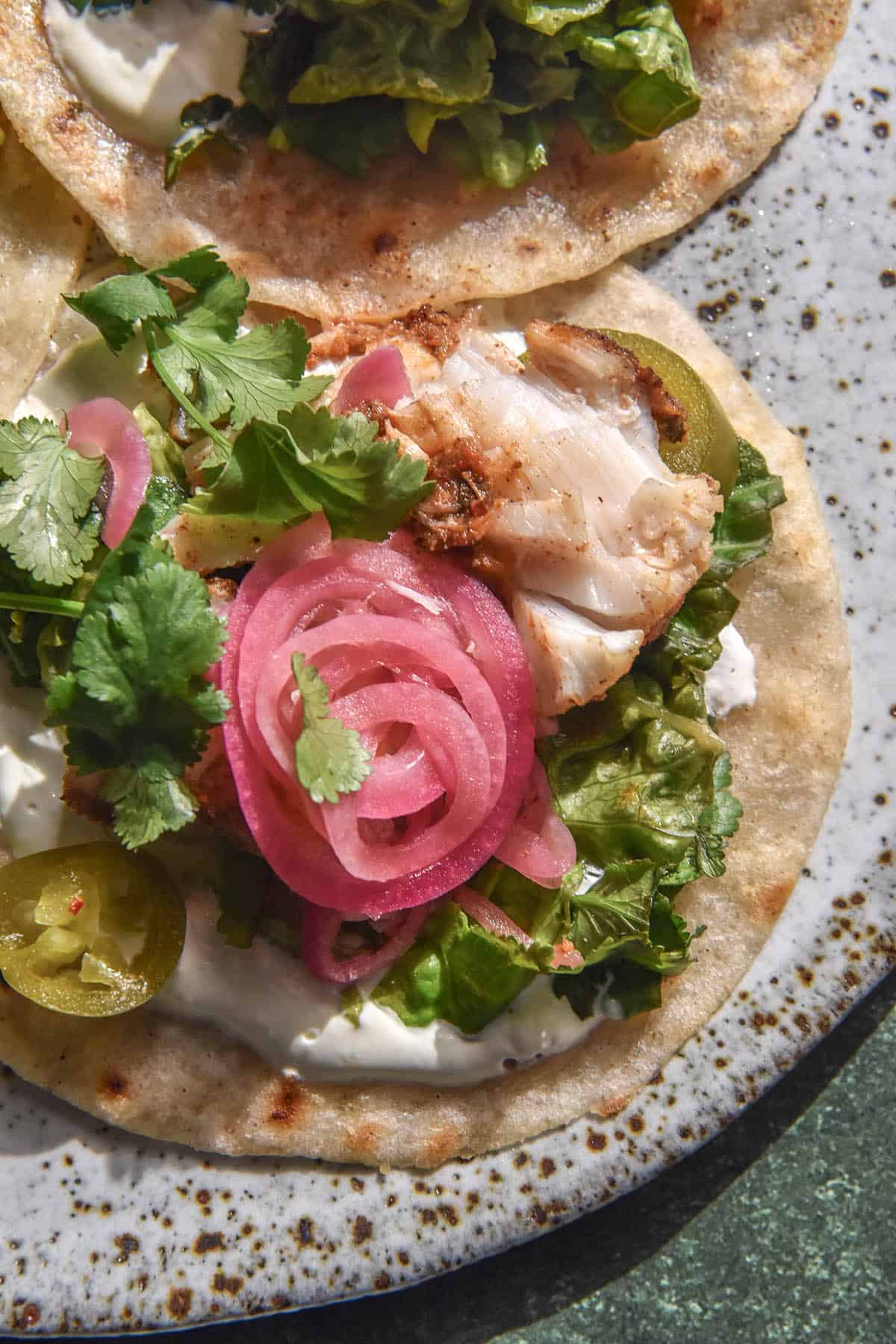 An aerial close up image of a low FODMAP fish taco topped with low FODMAP pickled onions, coriander and pickled jalapeño. The tacos sit atop a white speckled ceramic plate against a medium grey backdrop