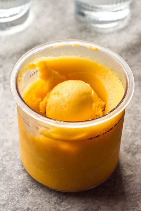 A side of brightly lit image of a tub of Mango and kombucha Ninja Creami on a concrete backdrop. Two glasses of water sit in the back of the image.