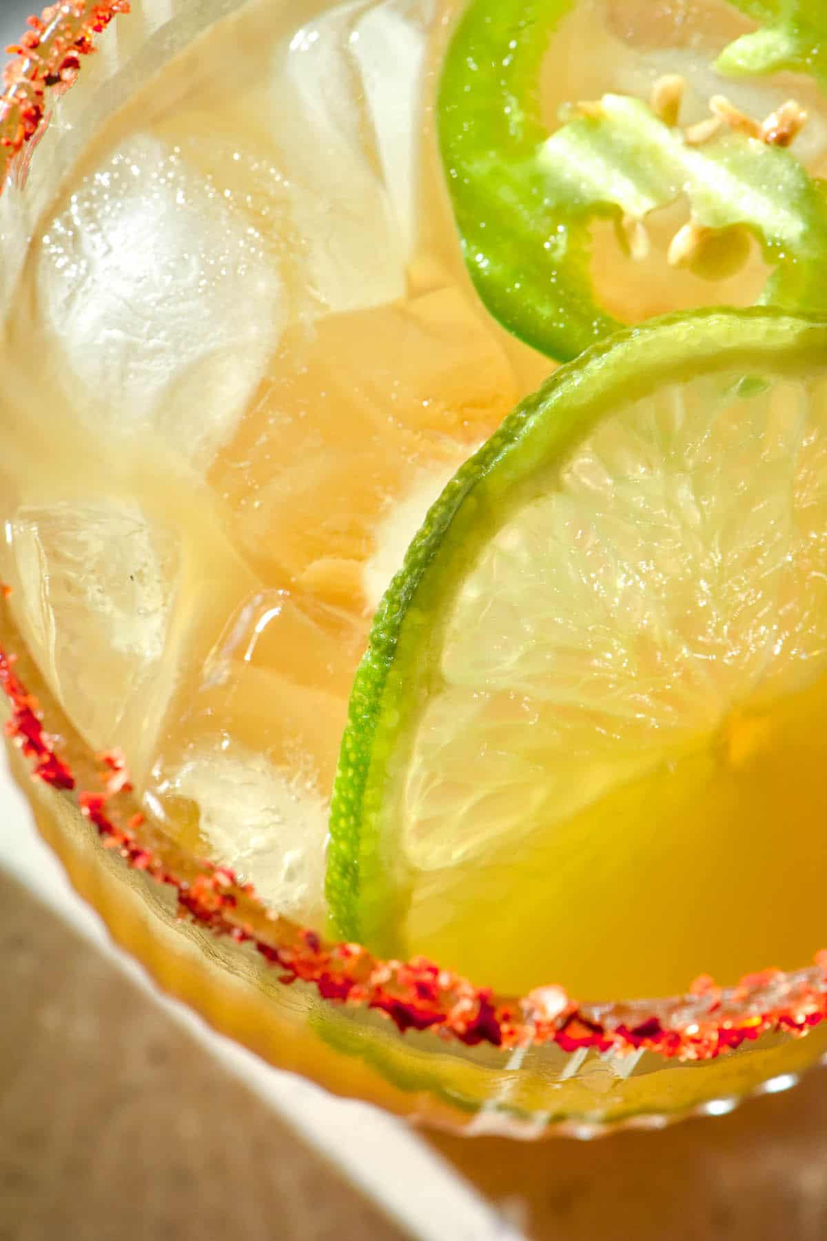 An aerial macro image of a cocktail margarita with a tajin rim, ice, slices of lemon and jalapeno