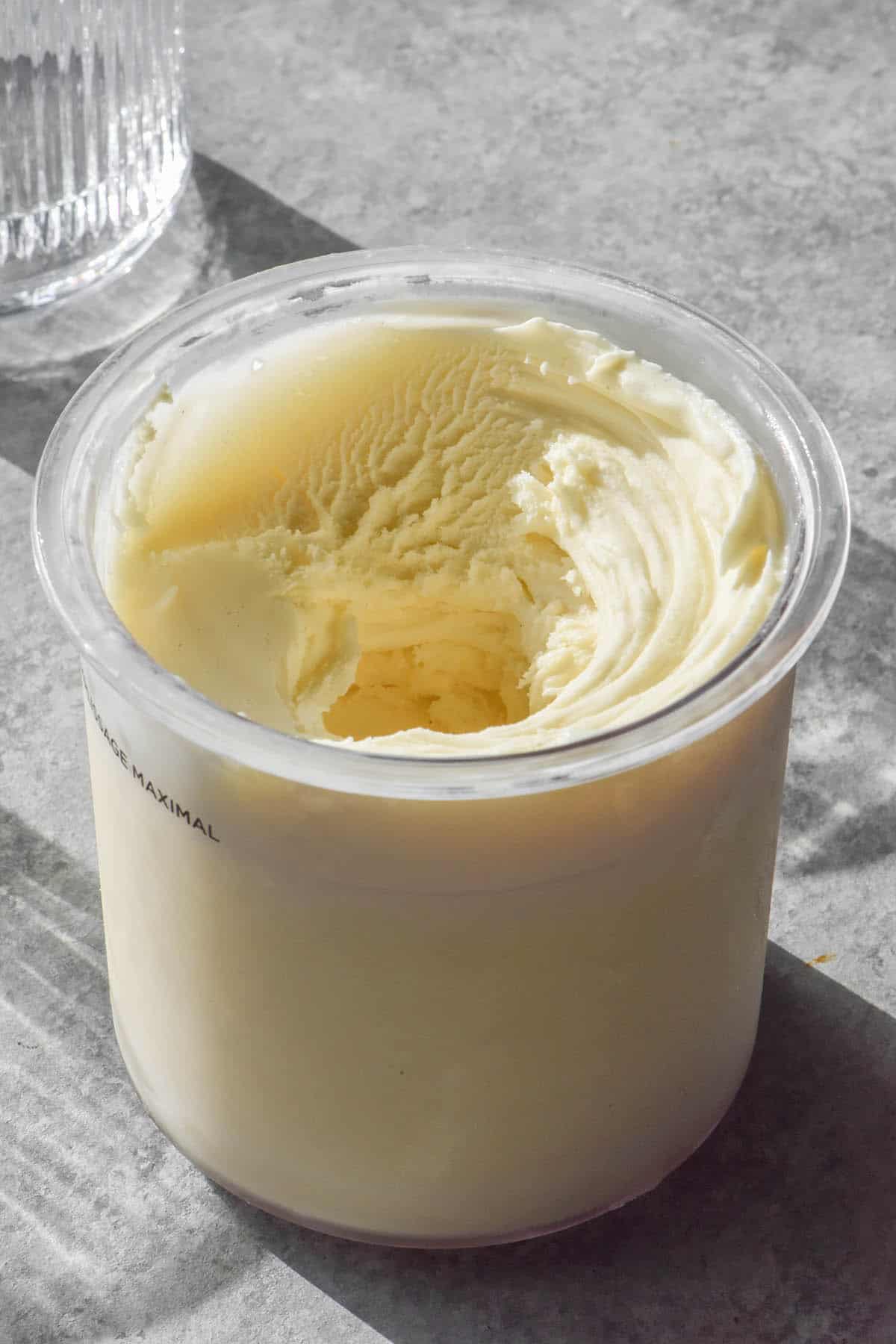 A side on image of a tub of Vanilla Ninja Creami on a concrete backdrop. Sunlit glasses of water sit to the top left of the image