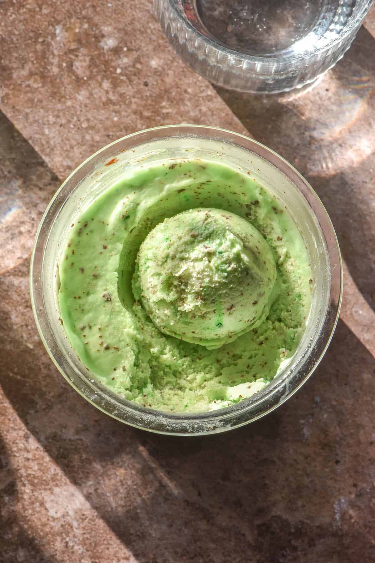 An aerial image of a tub of Ninja Creami choc mint ice cream atop a medium brown backdrop. Two sunlit glasses of water sit on either side of the ice cream tub.