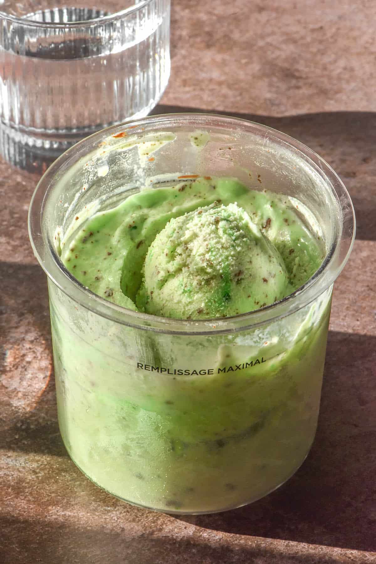 A side on image of a tub of green mint chocolate chip Ninja Creami ice cream on a light brown backdrop. 