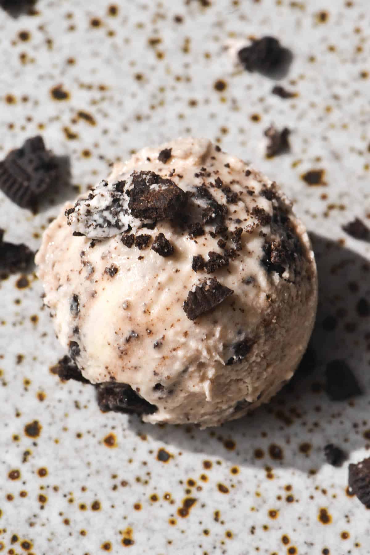 An aerial image of a scoop of cookies and cream ice cream on a speckled white ceramic plate. The scoop is topped with crumbled cookies.