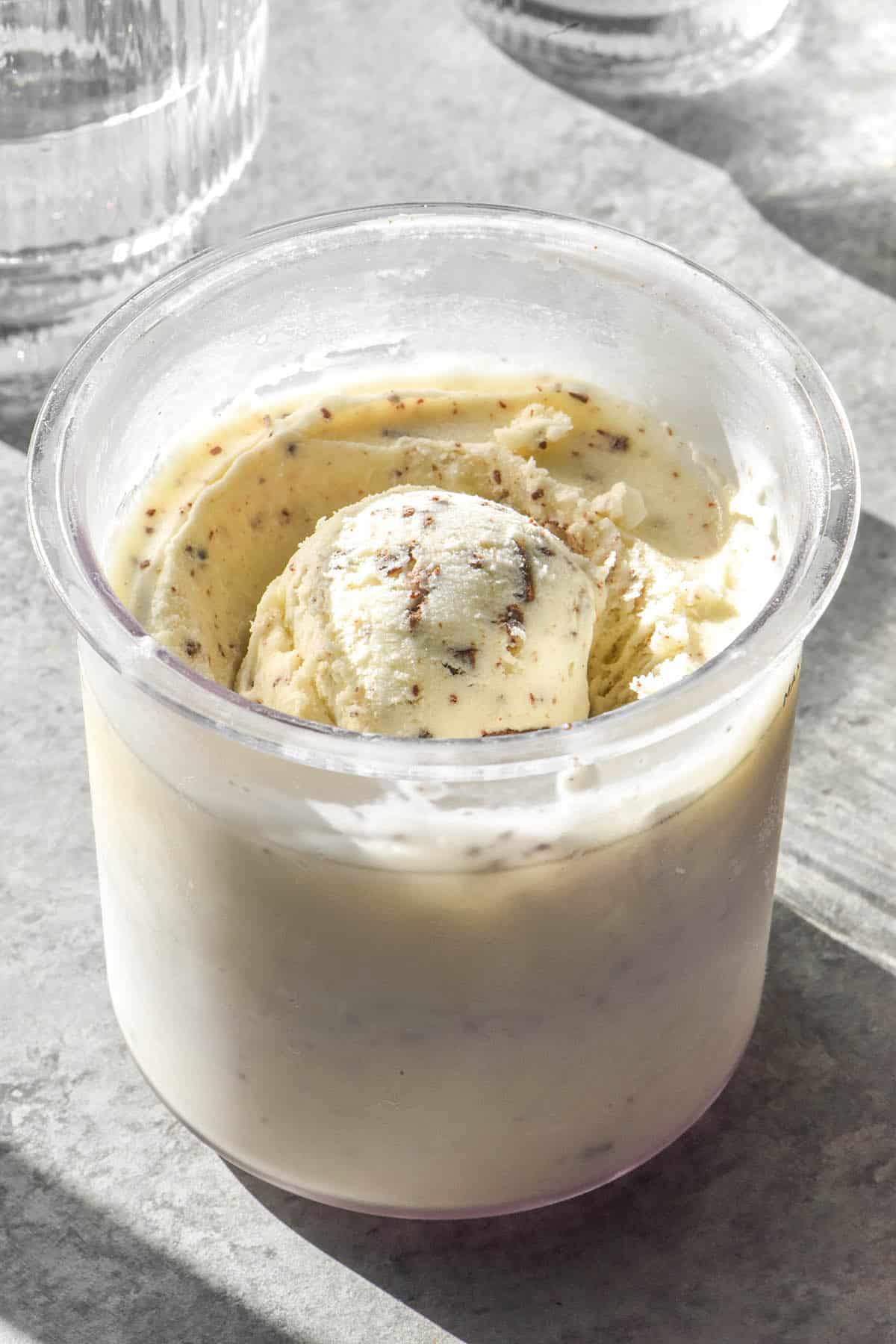 A side on brightly lit image of a tub of Ninja Creami mint chocolate chip ice cream on a concrete backdrop. The ice cream is flanked by two sunlit glasses of water 