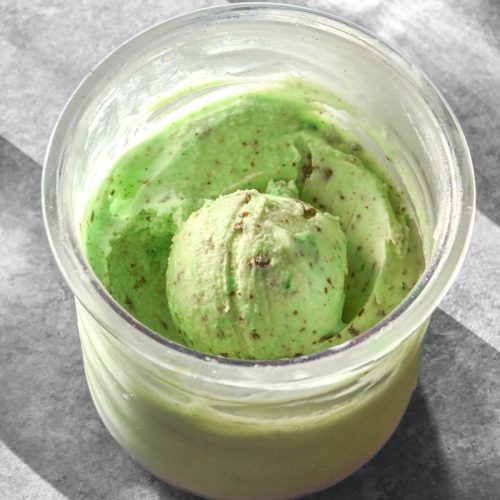 A sunlit aerial image of a tub of Ninja Creami mint chocolate chip ice cream with green food colouring. The ice cream sits on a light concrete backdrop and is flanked by sunlit glasses of water