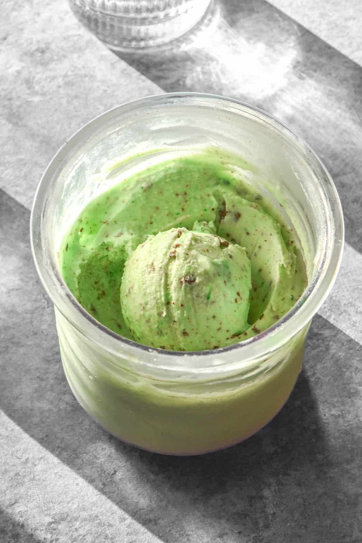 A sunlit aerial image of a tub of Ninja Creami mint chocolate chip ice cream with green food colouring. The ice cream sits on a light concrete backdrop and is flanked by sunlit glasses of water