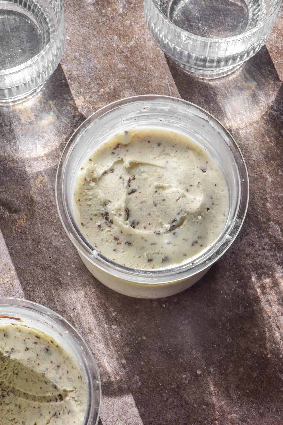 An aerial image of two tubs of Ninja Creami mint chocolate chip ice cream on a light brown backdrop. The ice cream tubs are flanked by two sunlit glasses of water.