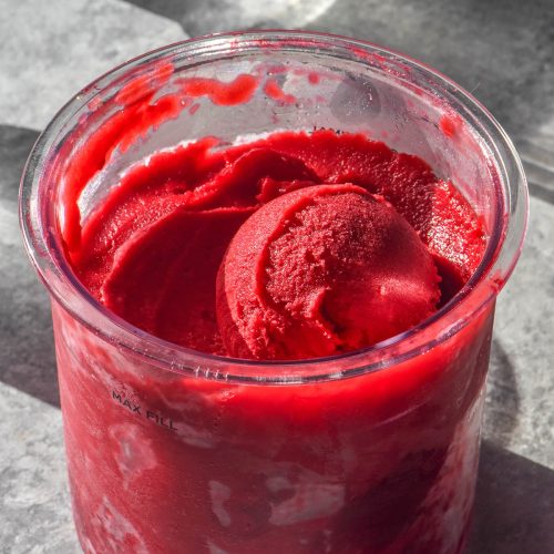 A side on brightly lit image of a Ninja Creami tub filled with raspberry sorbet. The tub is flanked by two sunlit water glasses and sits on a concrete backdrop