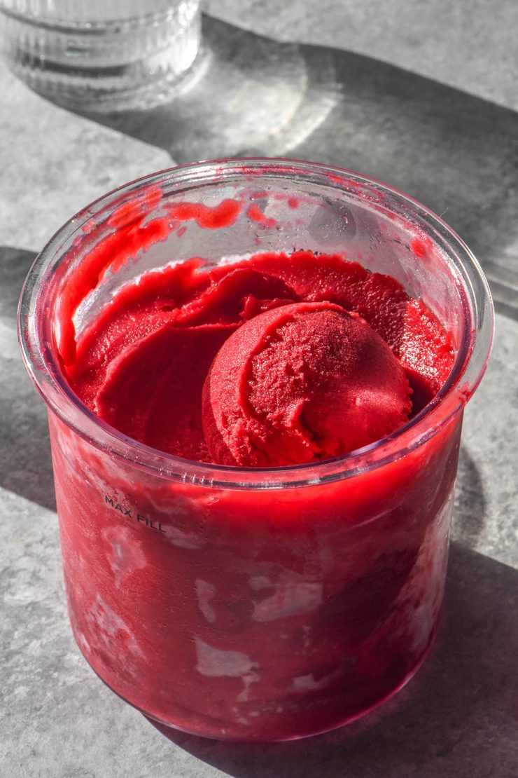 A side on brightly lit image of a Ninja Creami tub filled with raspberry sorbet. The tub is flanked by two sunlit water glasses and sits on a concrete backdrop