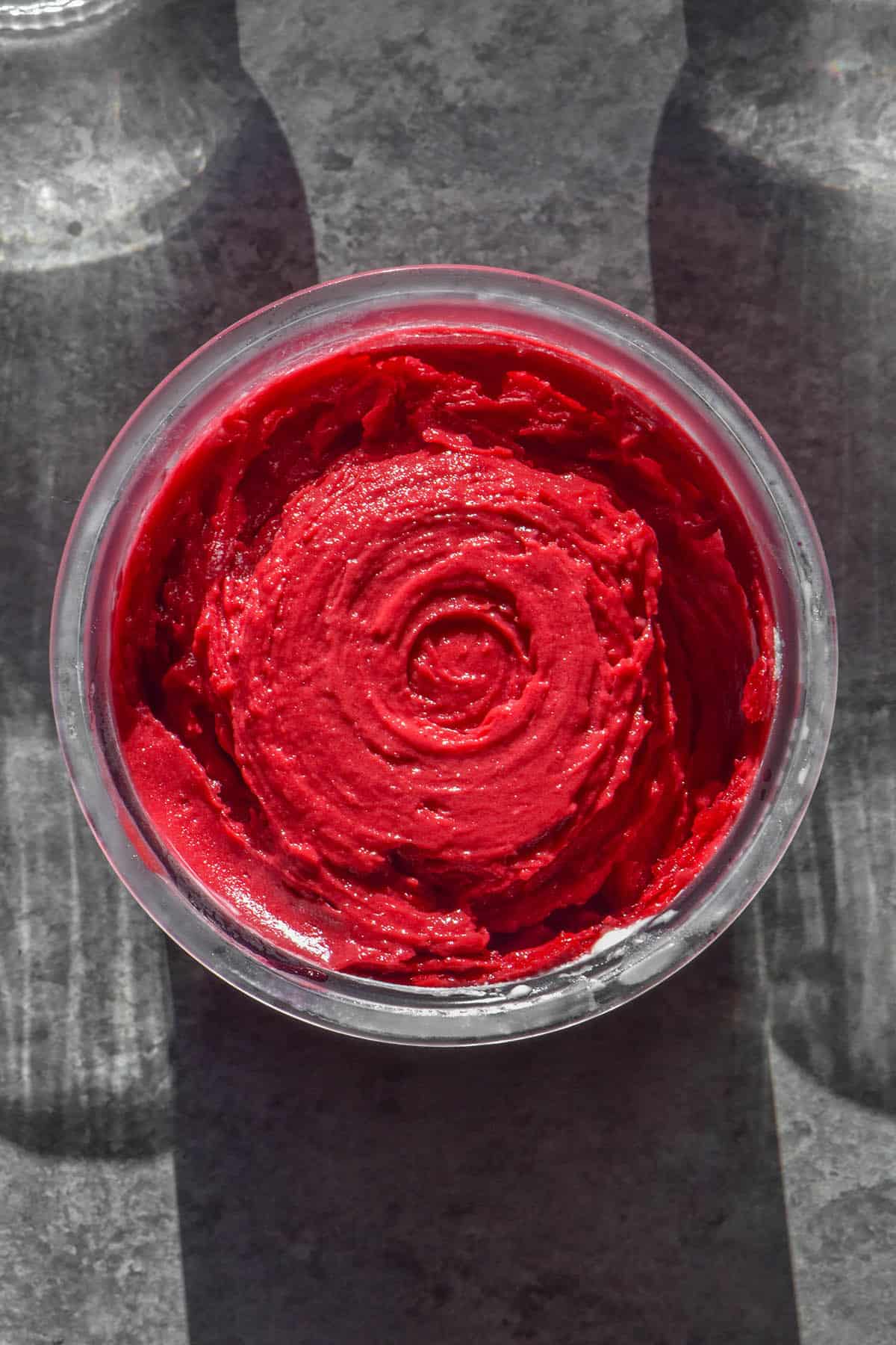 An aerial image of a tub of Ninja creami raspberry sorbet on a concrete backdrop. The tub is flanked by two sunlit glasses of water.