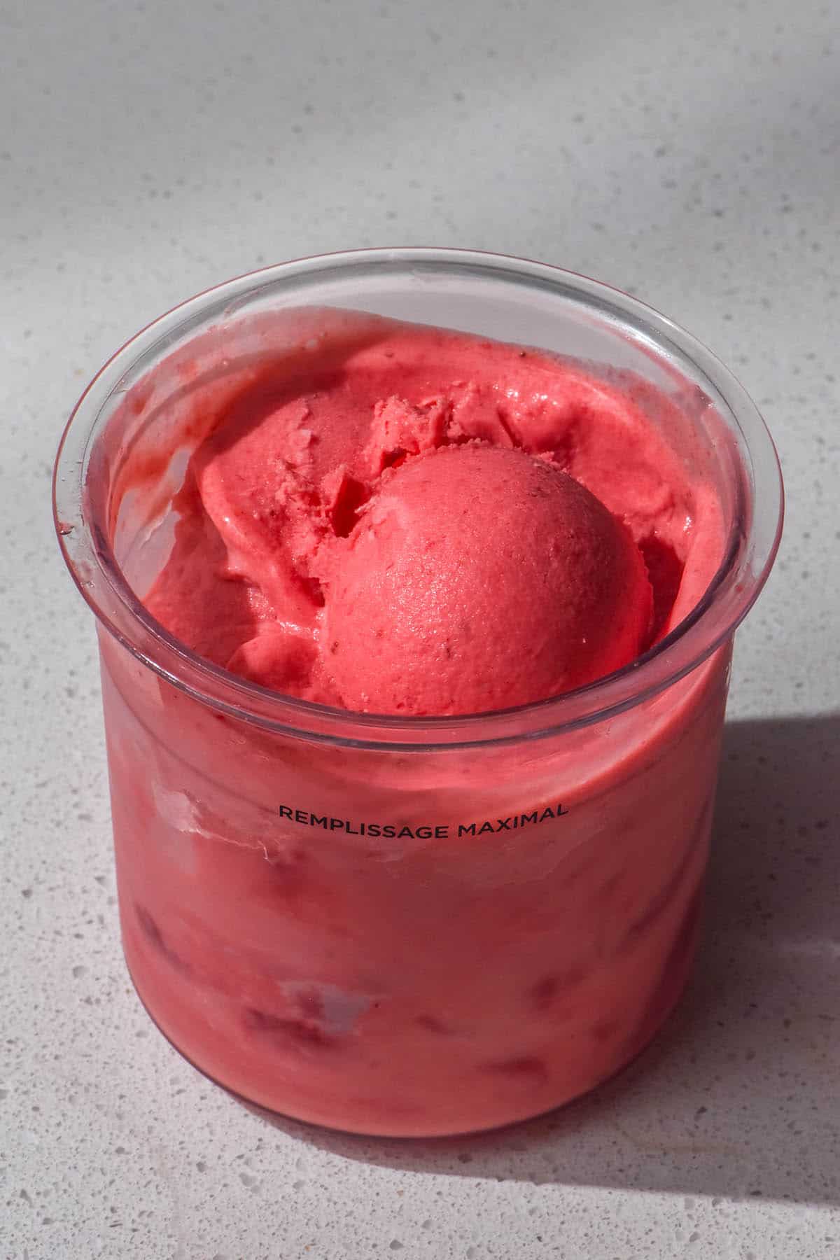 An aerial image of a tub of Ninja Creami strawberry ice cream on a white stone bench top. The ice cream tub has a scoop of the ice cream sitting atop the ice cream