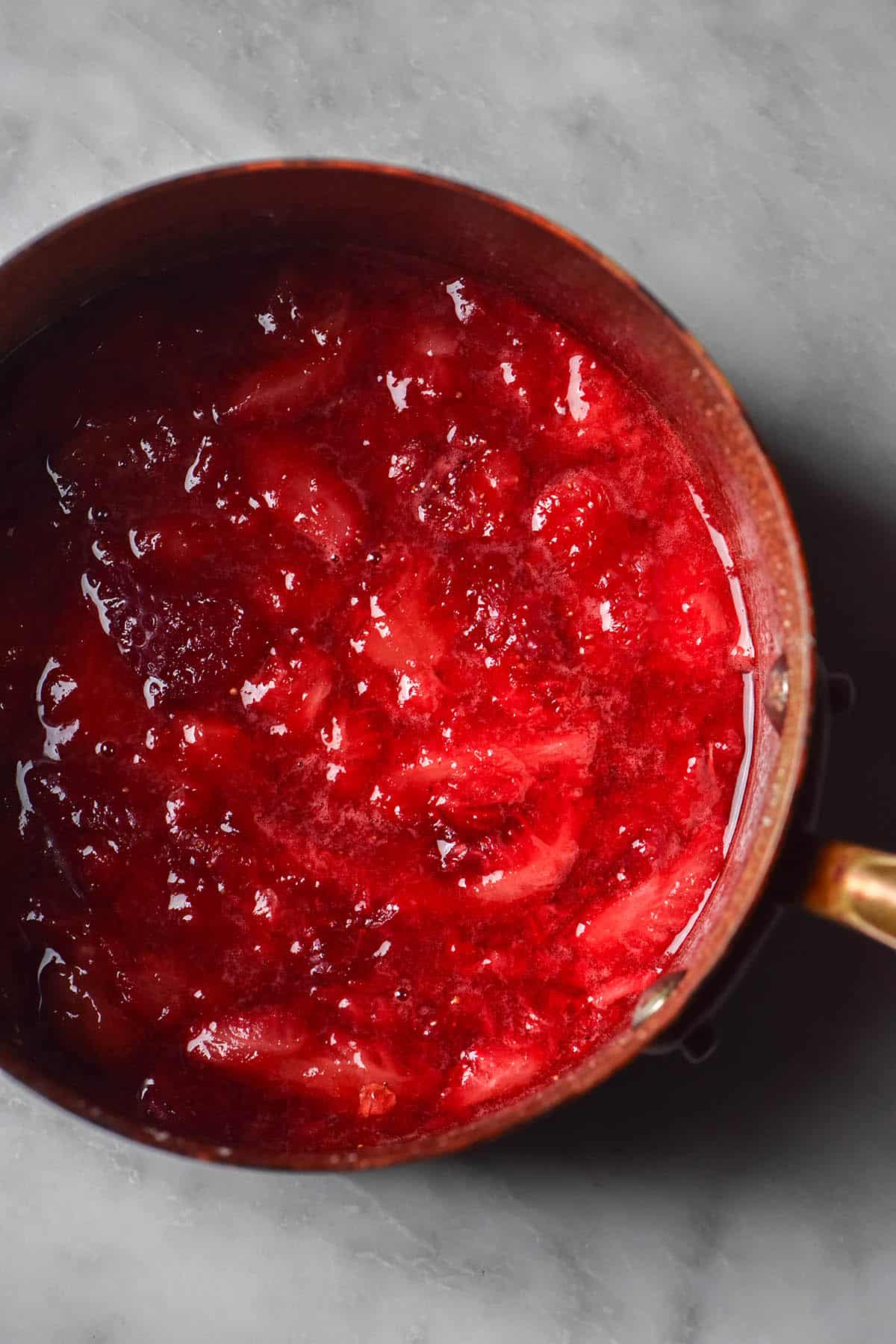 An aerial image of stewed strawberries and sugar in a small copper saucepan atop a white marble table