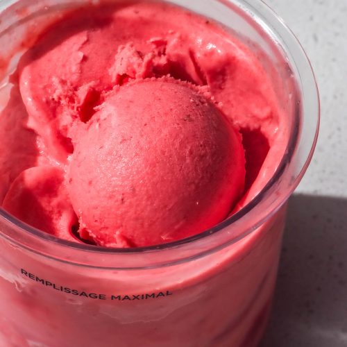 An aerial image of a tub of Ninja Creami strawberry ice cream on a white stone bench top. The ice cream tub has a scoop of the ice cream sitting atop the ice cream