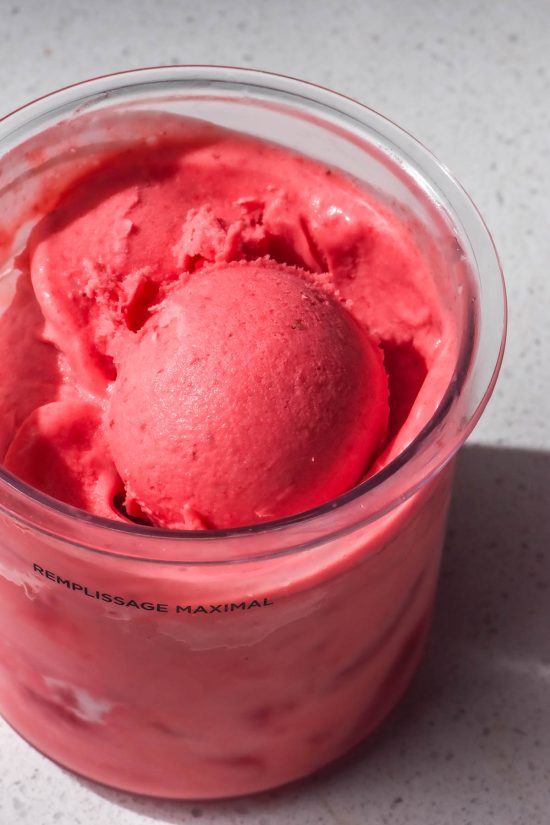 An aerial image of a tub of Ninja Creami strawberry ice cream on a white stone bench top. The ice cream tub has a scoop of the ice cream sitting atop the ice cream