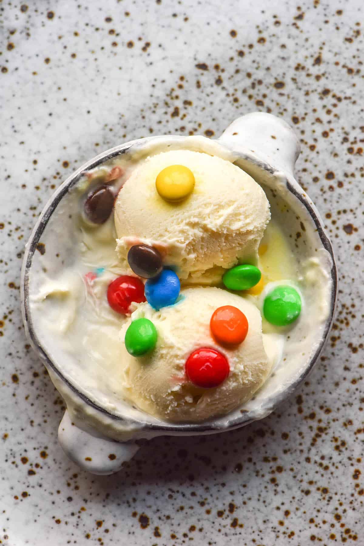 An aerial image of two scoops of vanilla ice cream in a small white ceramic bowl atop a white ceramic plate. The scoops are melting and topped with mnms
