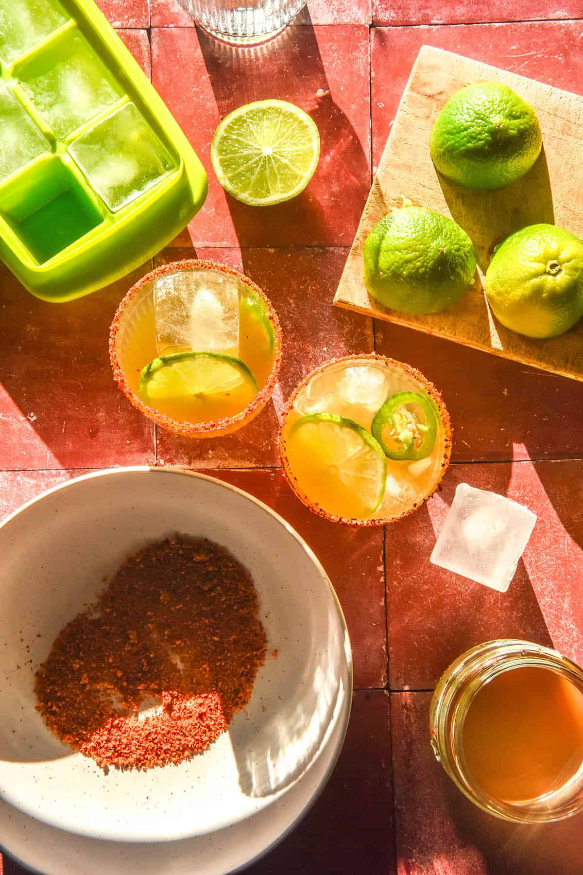 An aerial image of non alcoholic margaritas on a terracotta tile backdrop in bright sunlight. The margaritas are surrounded by extra limes, tajin and ice as well as a jar of non alcoholic margarita syrup.