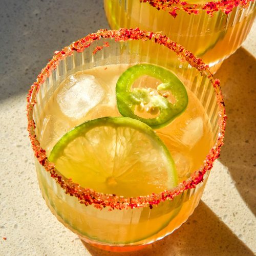 An aerial brightly lit image of two non alcoholic margaritas on a white stone bench top in bright sunlight. The margaritas have a slice of lime and jalapeño in each and both have a tajin salt rim