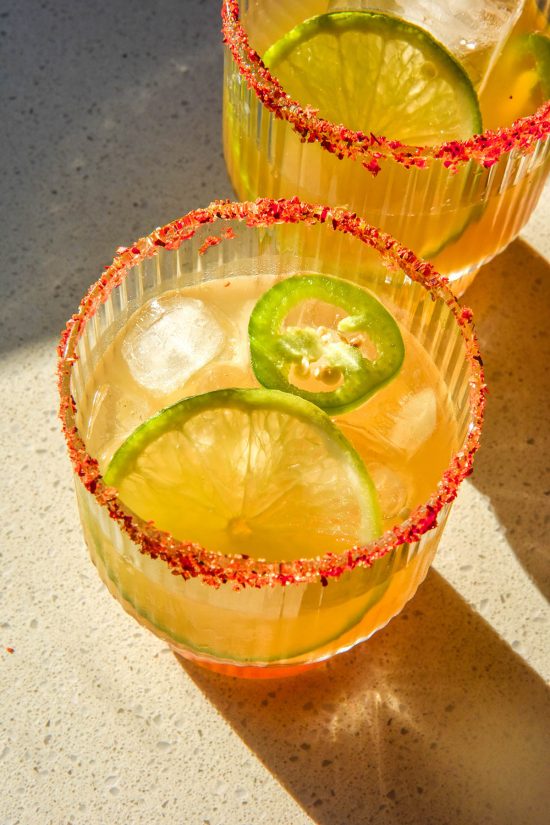 An aerial brightly lit image of two non alcoholic margaritas on a white stone bench top in bright sunlight. The margaritas have a slice of lime and jalapeño in each and both have a tajin salt rim