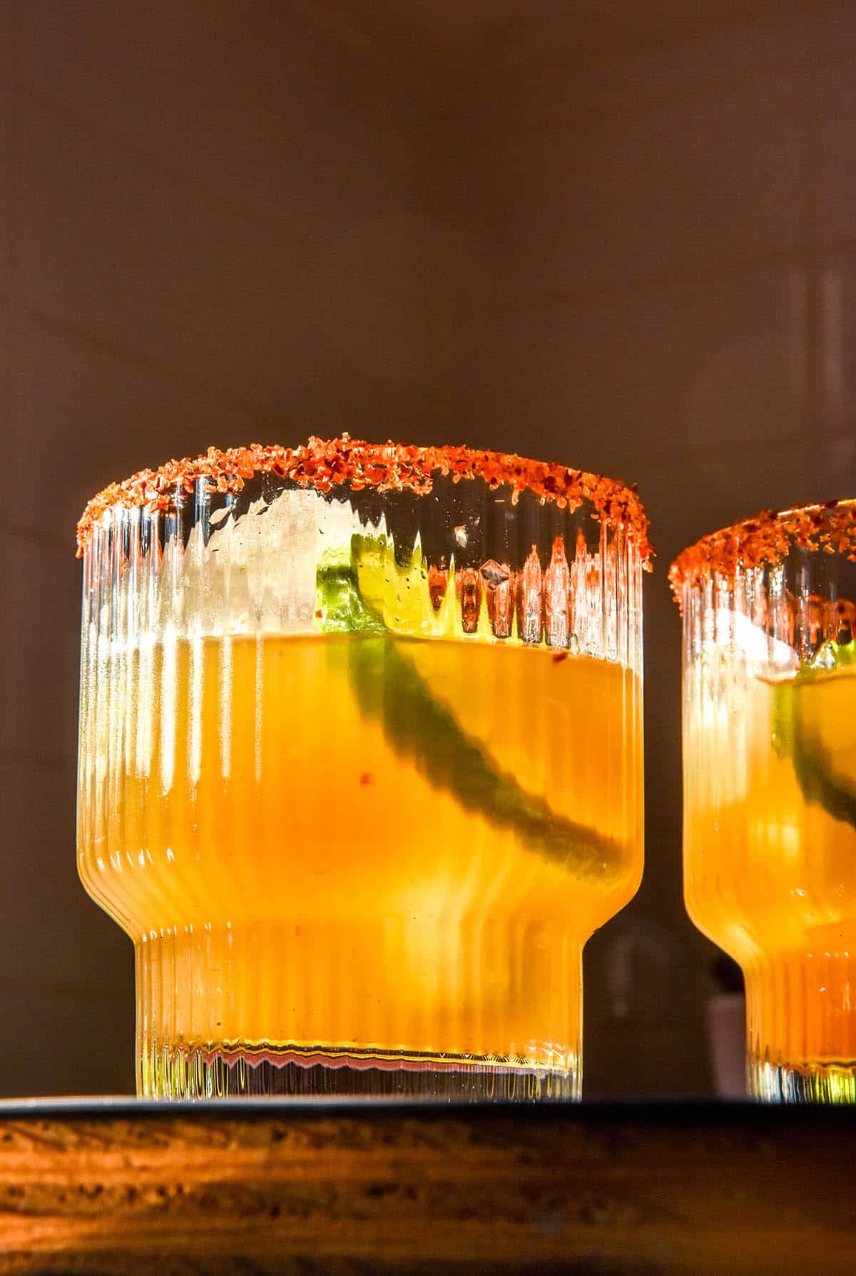 A moody image of two non alcoholic margaritas on a wooden table. The margaritas are in textured glasses and each has a tajin salt rim. The glasses are filled with ice, a slice of lime and a slice of jalapeño 