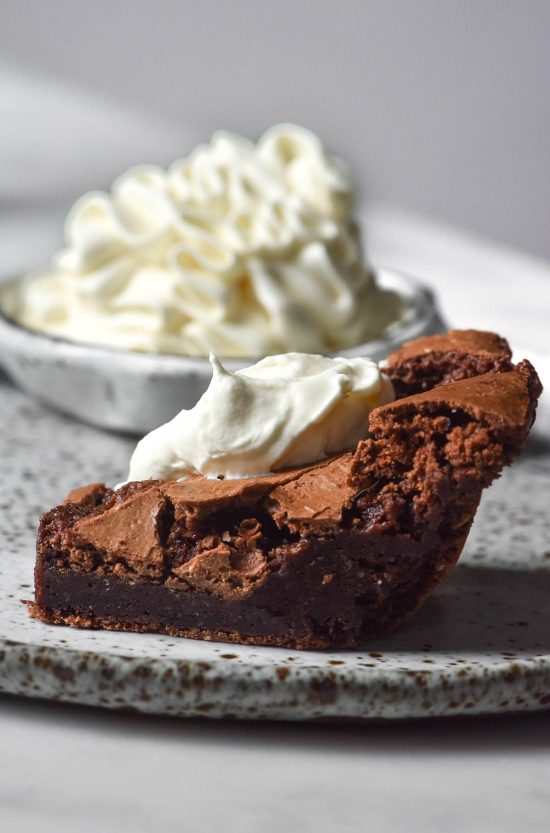 A side on image of a brownie topped with stabilised lactose free whipped cream. The brownie sits atop a white ceramic plate and a smaller bowl of whipped cream sits in the back of the image