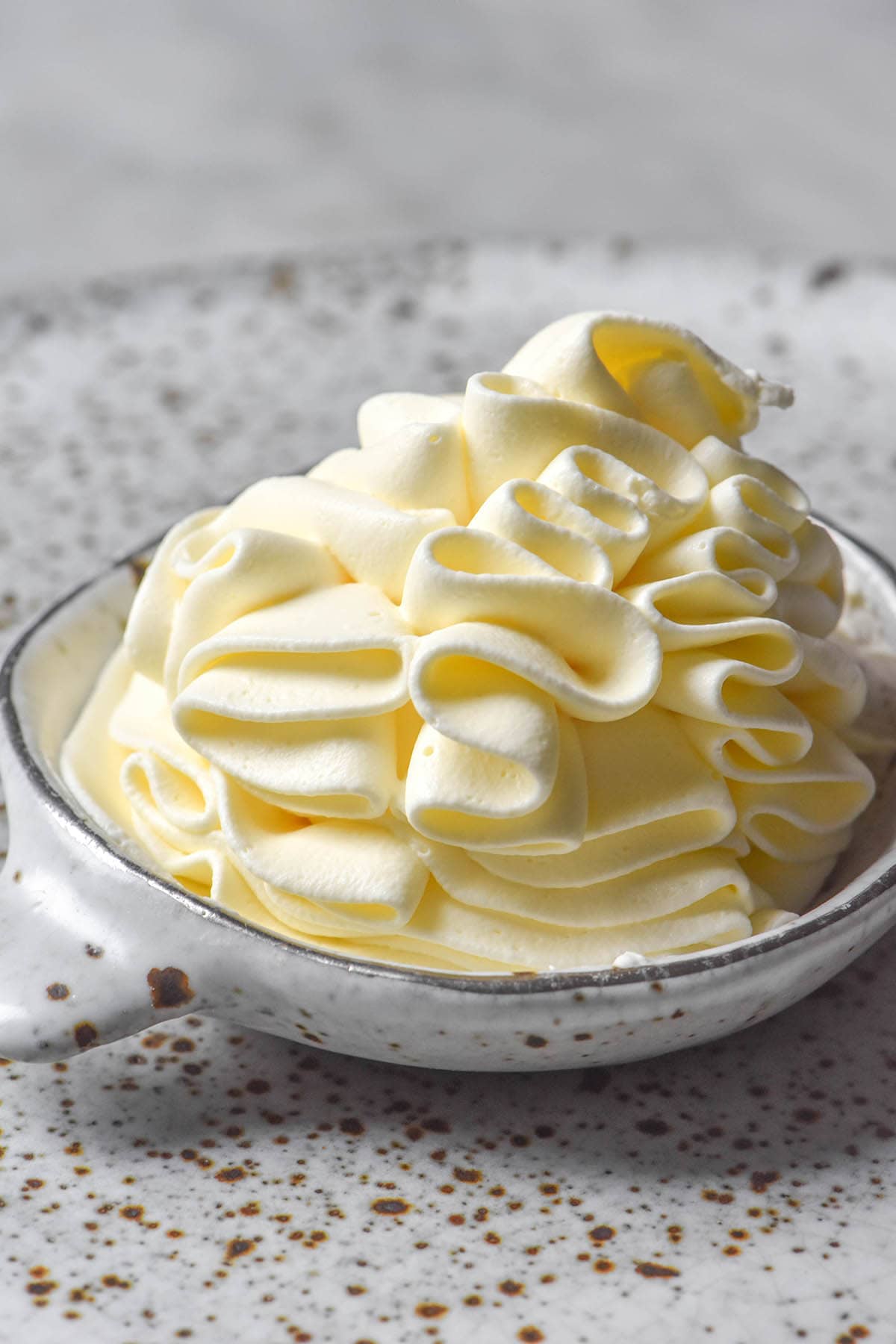 A side on image of a small white ceramic bowl filled with piped lactose free whipping cream. The bowl sits atop a white speckled ceramic plate