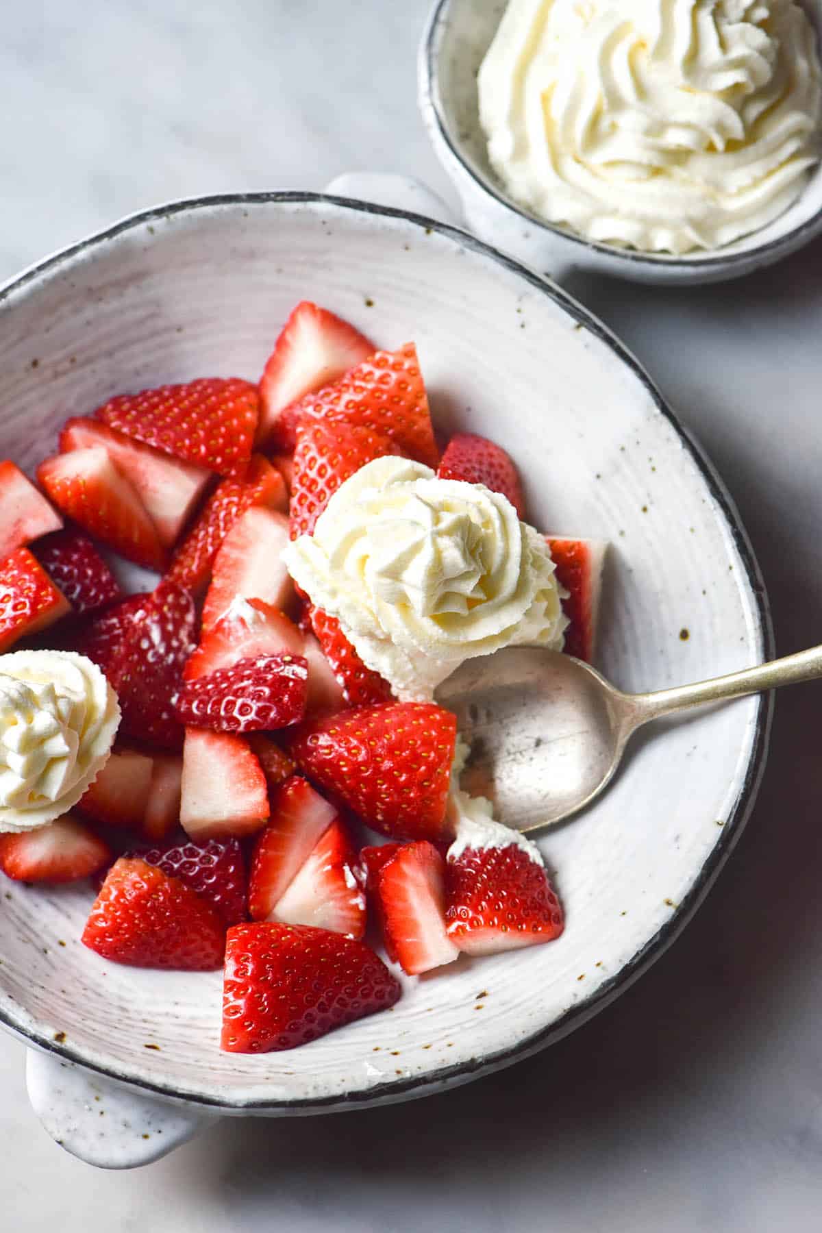 An aerial image of a white ceramic bowl filled with strawberries and lactose free whipped cream. The bowl sits atop a white marble table and another smaller bowl of whipped cream sits off to the right of the main bowl