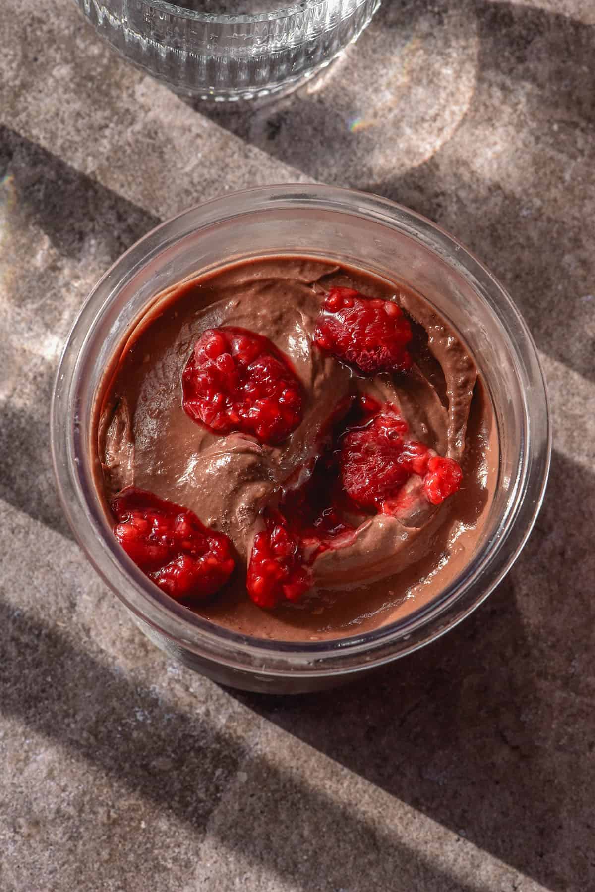 A sunlit aerial image of a tub of choc raspberry Ninja Creami on a dark grey backdrop. The ice cream is topped with extra raspberries