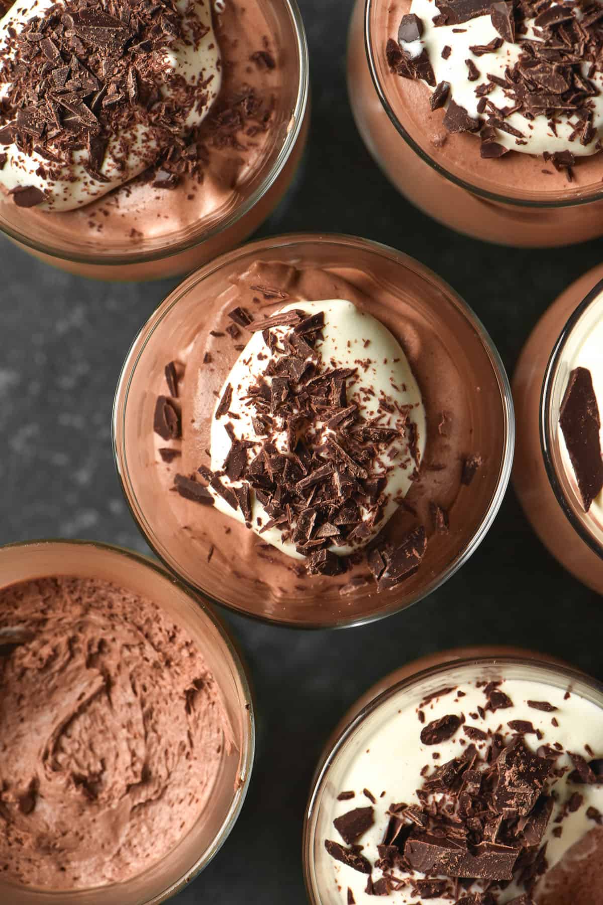 An aerial image of pots of dairy free chocolate mousse topped with dairy free whipped cream and chocolate shards against a dark grey backdrop