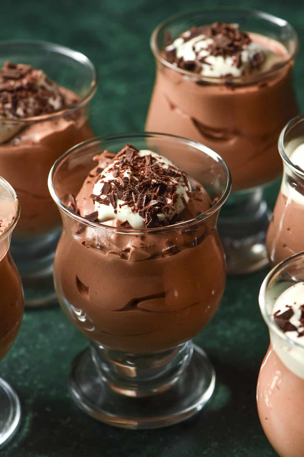 A side on image of serving glasses filled with dairy free chocolate mousse which is topped with dairy free whipped cream and chocolate shards. The glasses sits atop an olive green backdrop.