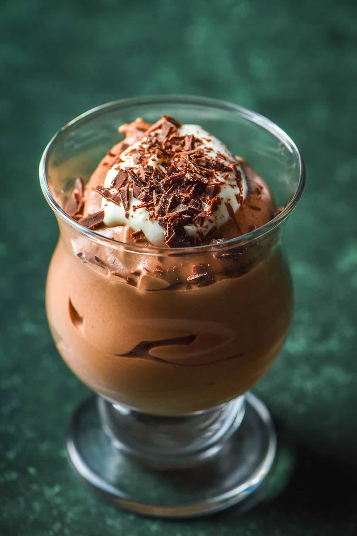 A side on image of a serving glass filled with dairy free chocolate mousse which is topped with dairy free whipped cream and chocolate shards. The glass sits atop an olive green backdrop. 