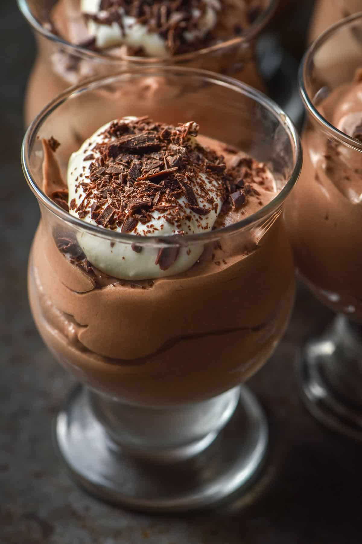 A close up side on image of pots of chocolate mousse topped with whipped cream and chocolate shards atop a dark grey backdrop