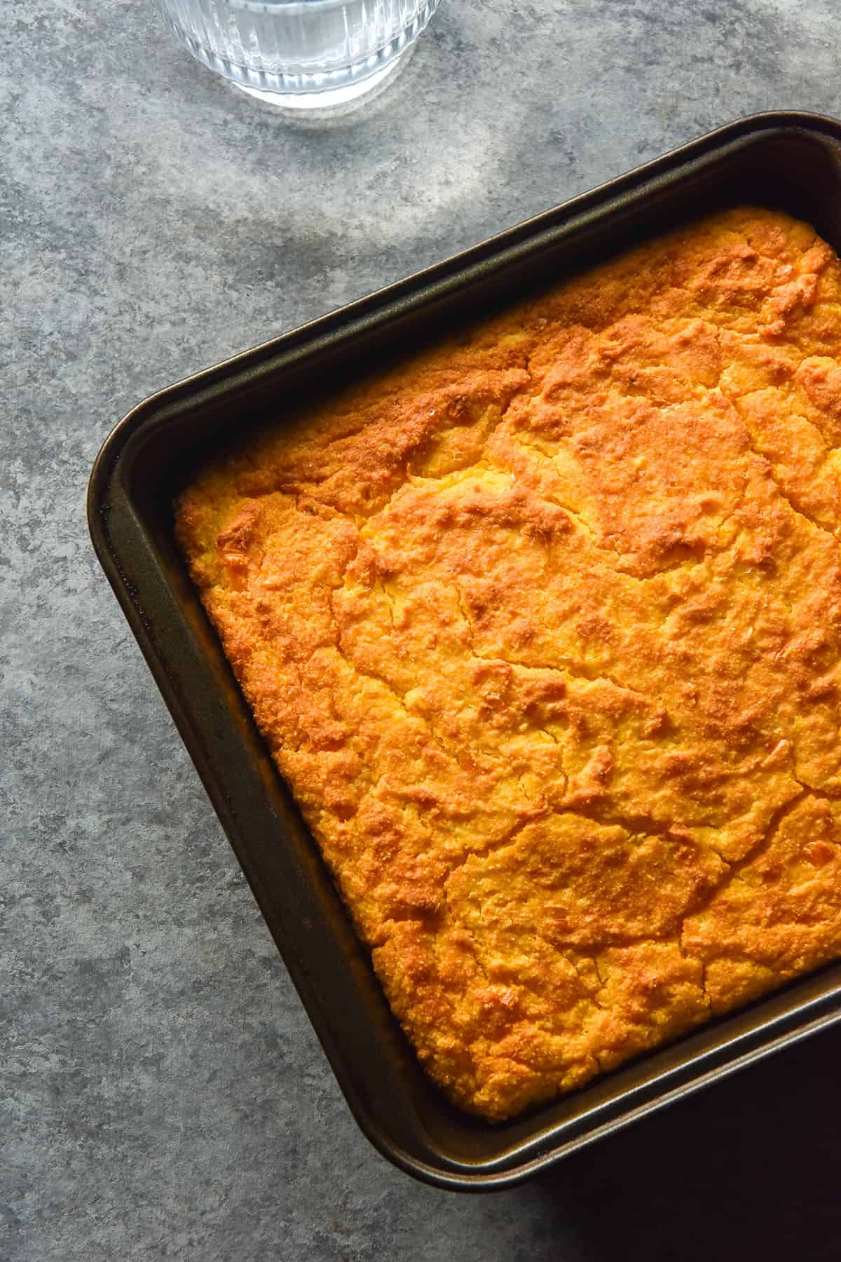 An aerial image of a square baking tin filled with gluten free cornbread on a light blue backdrop. 